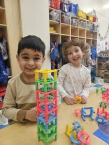 A young boy and a girl smile at the camera as they build with plastics blocks at their classroom table