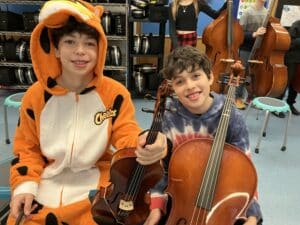 two seated boys smile as one boy, dressed in a tiger costume, holds a violin,and another boy holds a cello