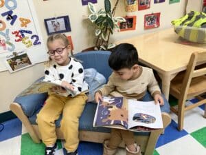 A boy and a girl in pajamas read side by side seated on t a sofa in their classroom