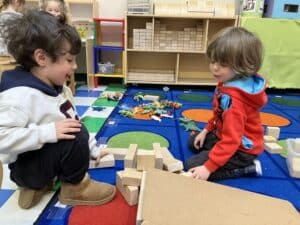 Two little boys smiling as they are seated on a colorful rug in their classroom while they build a structure with wooden blocks