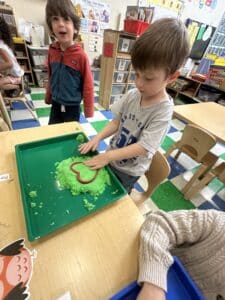 A boy cuts out a heart from green playdoh on a green tray as his classmate smiles for the photographer