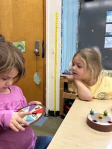 Two little girls seated at a classroom table. One watches as the other spins a dial