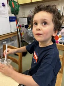 A young student seated at a table look sup at the camera as he tastes applesauce his class made