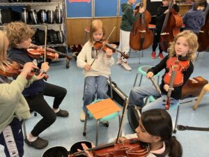 A group of five students play orchestral string instruments seated in a circle in their classroom