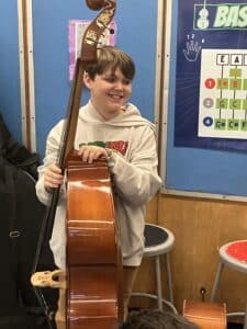 A boy smiles as he stands by a double bass in his music classroom